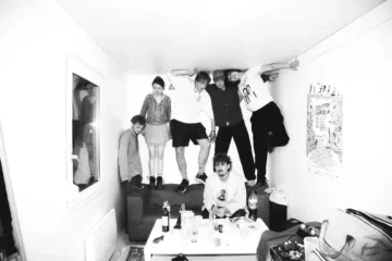 Fat Dog, black-and-white press photo of the six-piece band posed in a cluttered living room, several members perched on furniture and an exposed ceiling beam