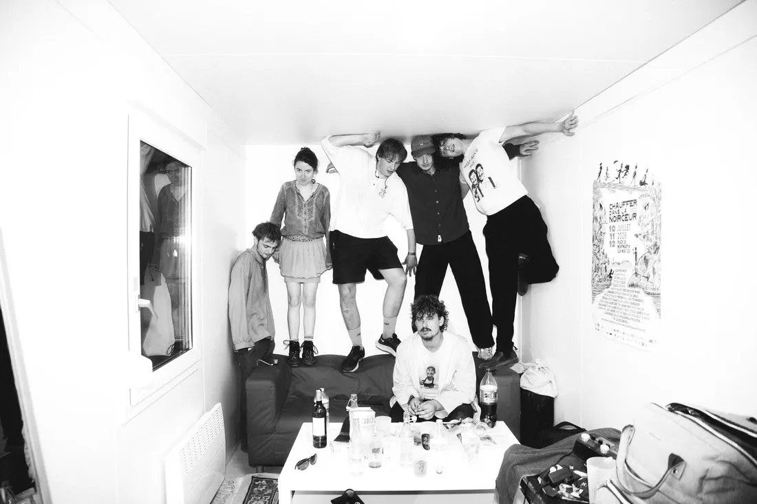 Fat Dog, black-and-white press photo of the six-piece band posed in a cluttered living room, several members perched on furniture and an exposed ceiling beam
